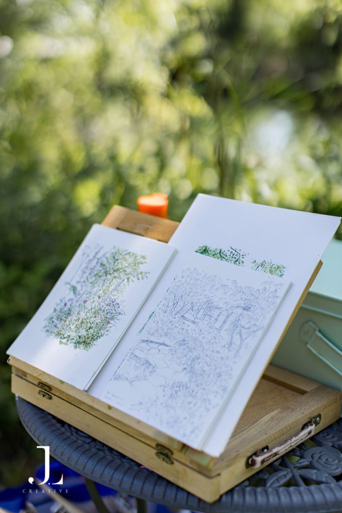 Open sketchbooks displaying watercolor landscape paintings on an easel outdoors.