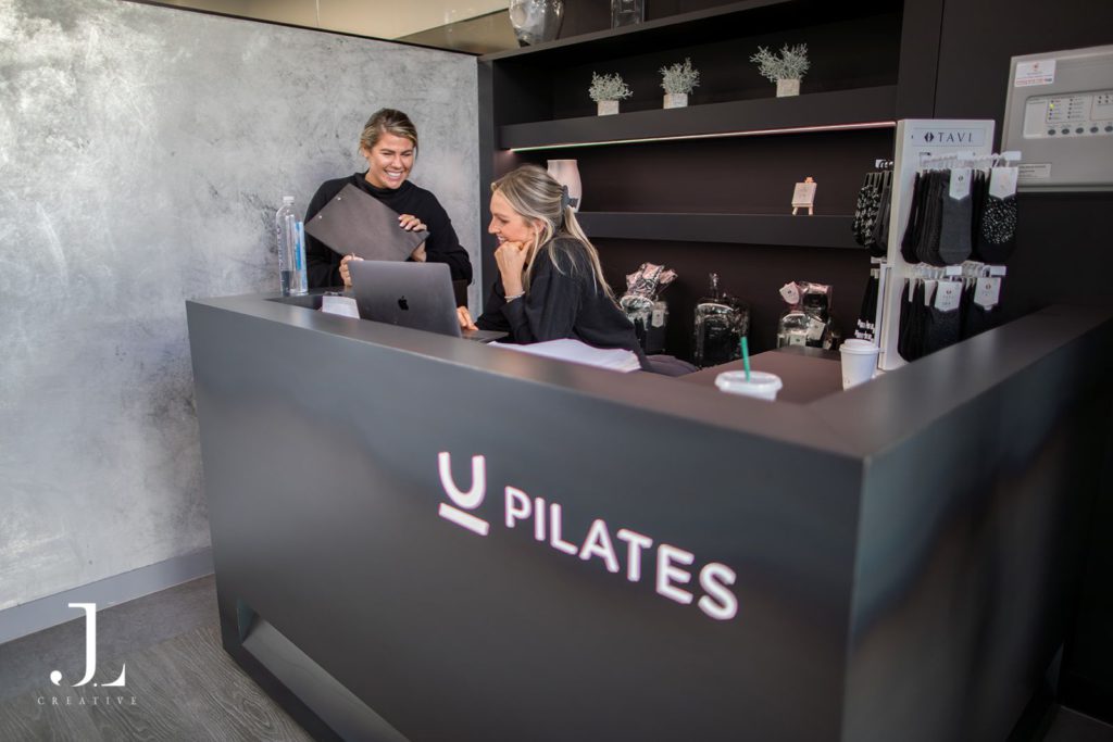Two staff members greeting clients behind the reception desk at You Pilates studio in Cobham.
